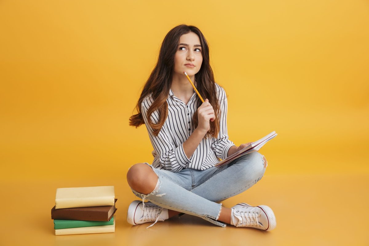 Adolescente sentada no chão aos lados de pilha de livros estudando com um caderno no colo e um lapis na mão enostando no queixo com expressão pensativa 