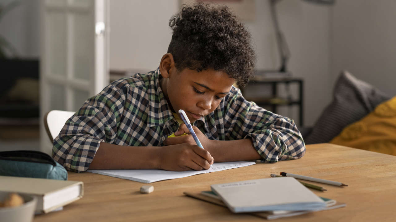 Estudante escrevendo em caderno com outros materiais de estudos sobre a mesa