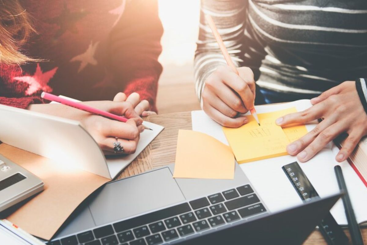 Dois estudantes na mesa escrevem em seus cadernos. É possível ver um notebook e alguns materiais espalhados na mesa