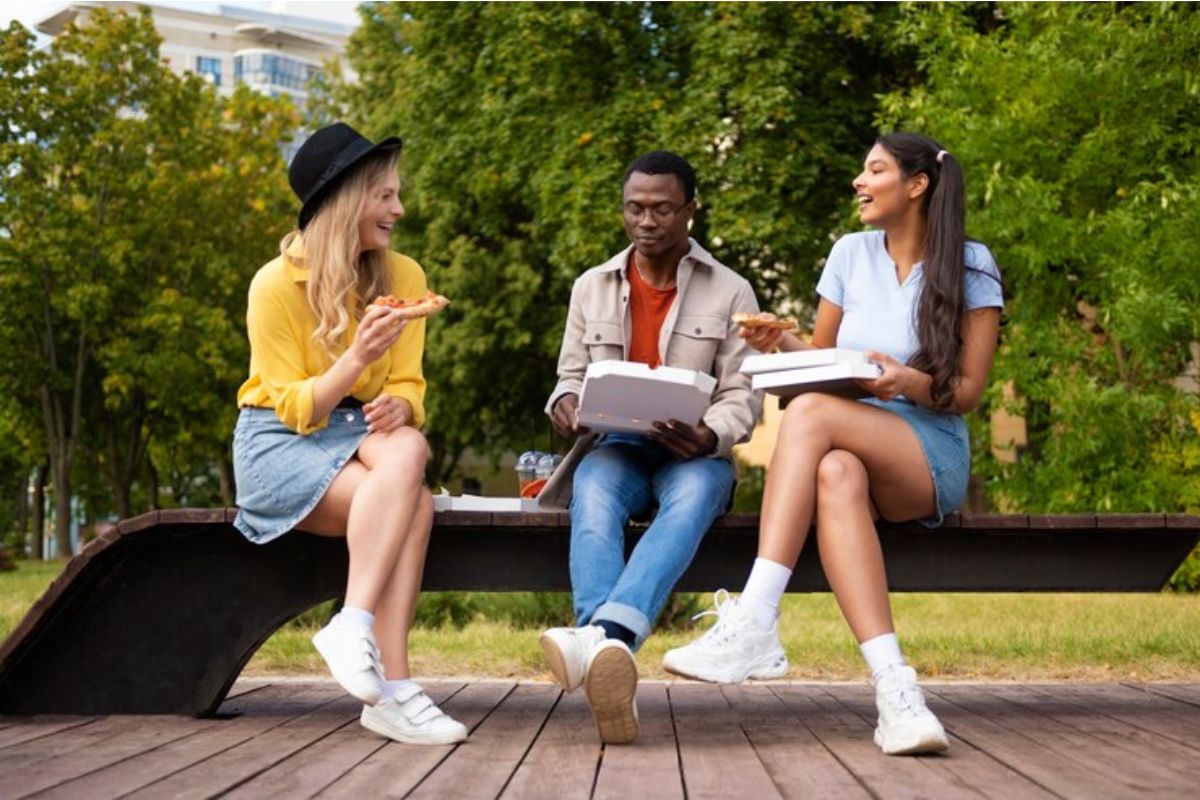 Três estudantes na praça da escola. Eles estão sentados em um banco e comem pizza. São duas garotas e um menino