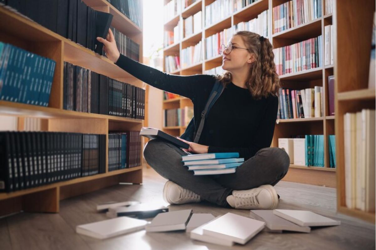 Uma menina sentada no chão da biblioteca estuda com um caderno no colo