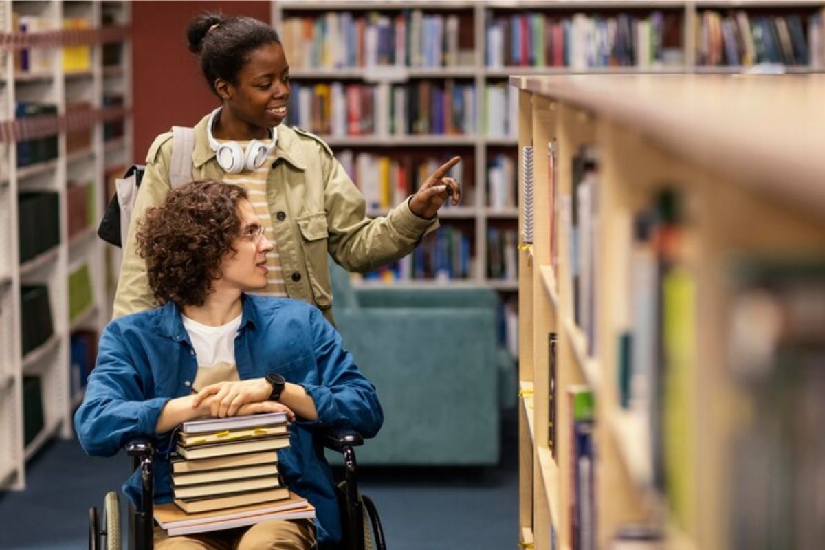 Dois amigos na biblioteca. Uma colega empurra seu amigo que está na cadeira de rodas. Eles escolhem livros na biblioteca