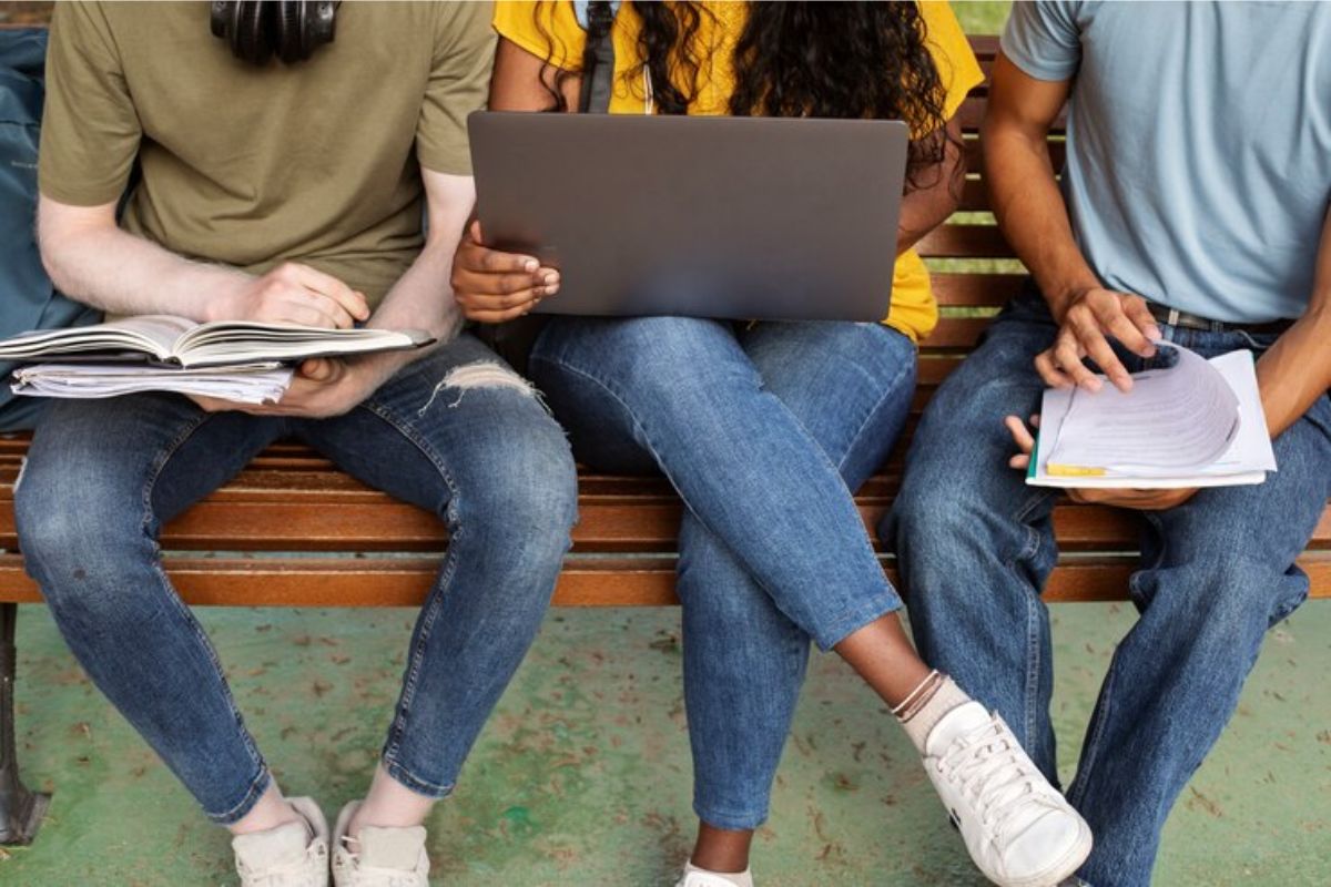 Três estudantes sentados em um banco. Dois estão com um caderno na mão, e uma aluno meio com o notebook confere os  Cursos MAIS Concorridos da ETEC 2025