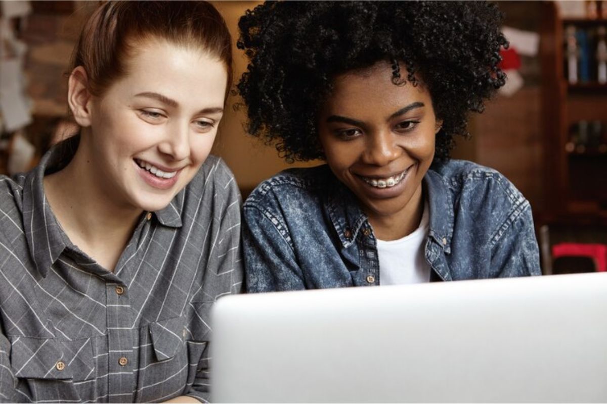 Duas estudantes conferem informações no notebook. Eles estão sentados em uma sala de estudos