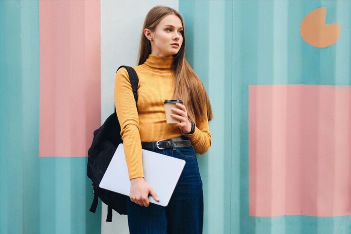 Uma estudante tira foto em frente a um muro colorido. Ela segura um copo de café em uma mão e seus materiais em outra