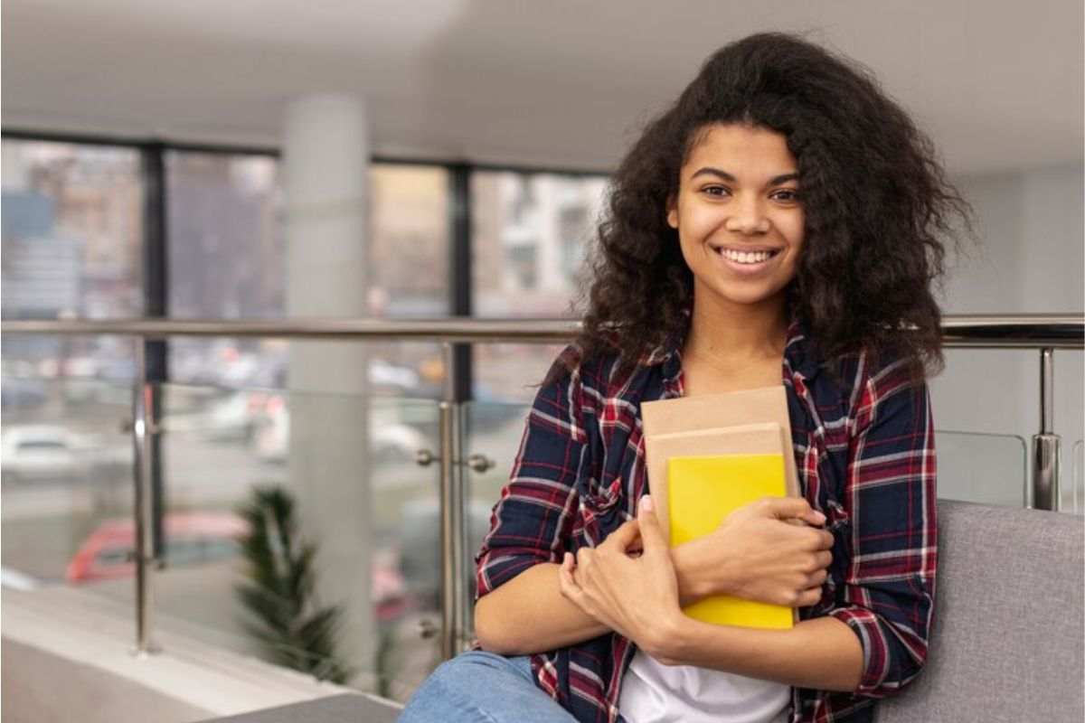 Uma estudante sorri para a foto e segura um caderno de capa amarela contra o colo