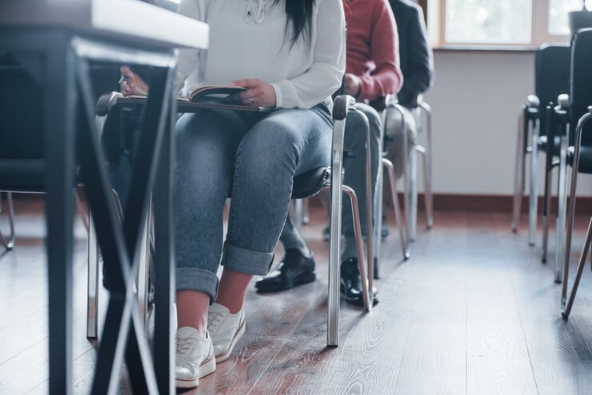 Imagem de uma sala de aula com estudantes sentados. A altura da foto pega apenas as pernas dos estudantes 