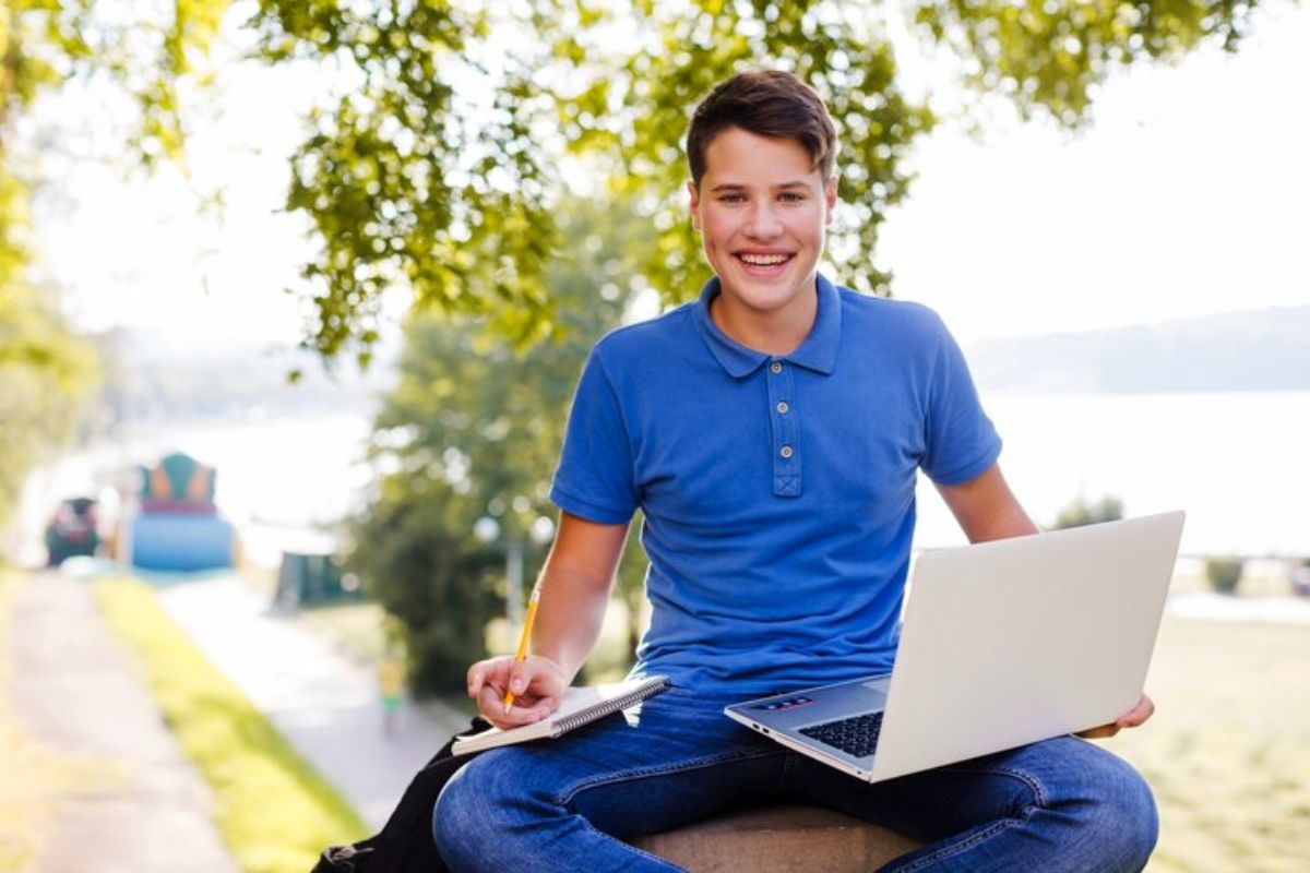 Um estudante na praça da escola com o notebook aberto em cima da perna e um caderno aberto na outra perna