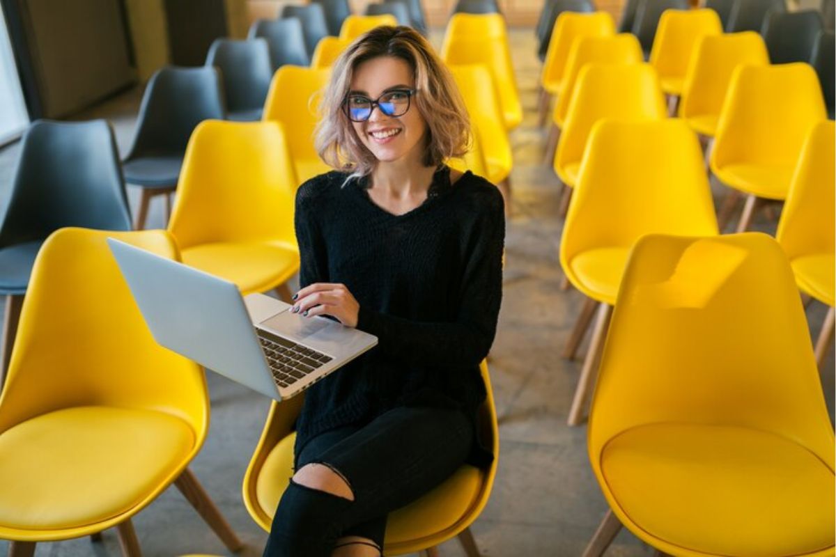 Uma aluna sentada em uma sala de eventos sozinha com seu notebook aberto na mão