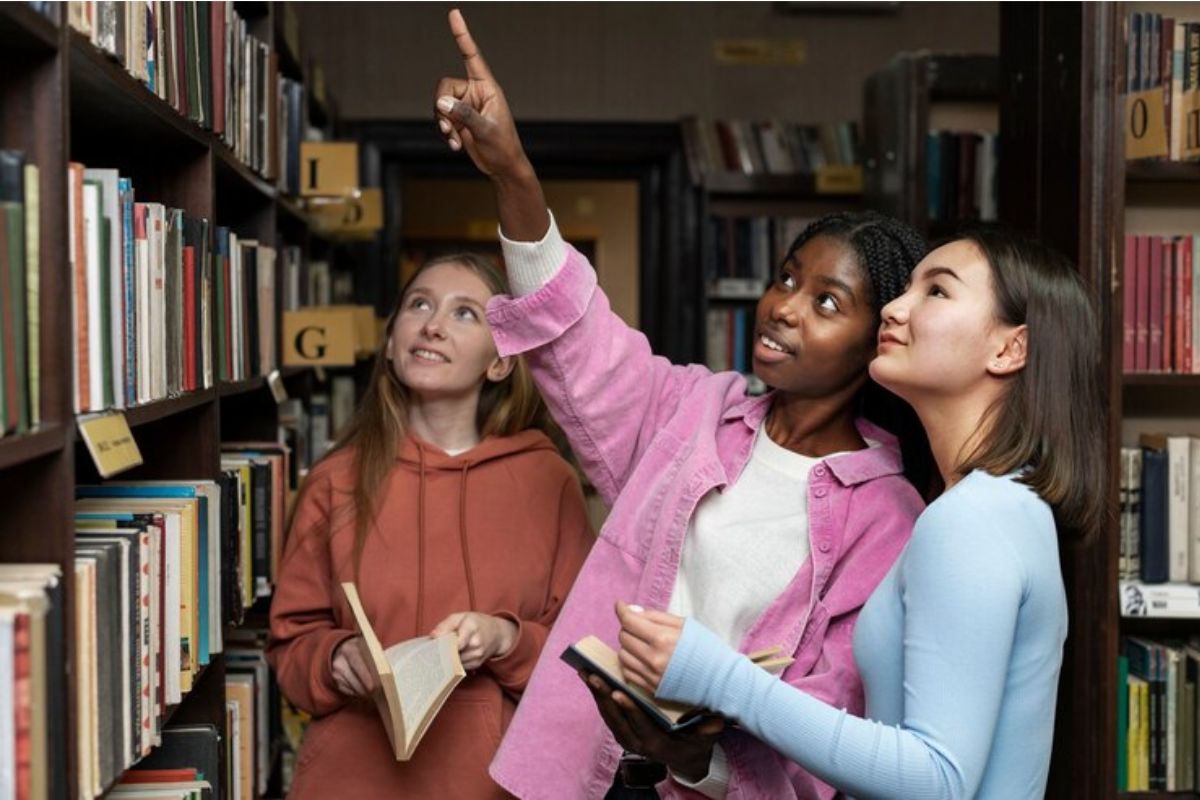 Três estudantes estão na biblioteca. Elas escolhem um livro na prateleira 