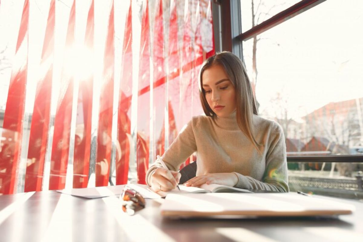 Uma aluna na sala de aula escreve em seu caderno apoiada na mesa. Ao fundo, o sol entra pela janela, com uma cortina vermelha