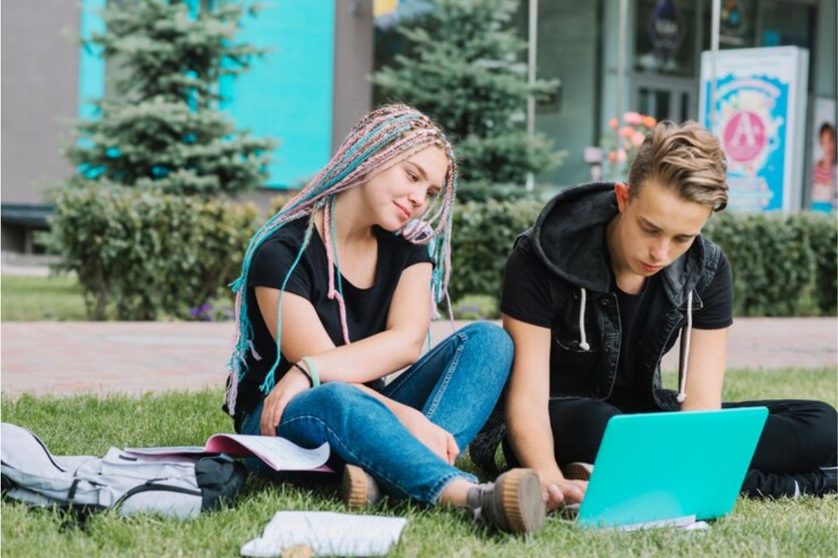 Dois estudantes sentadas no chão de grama da escola mexem no notebook para estudar 