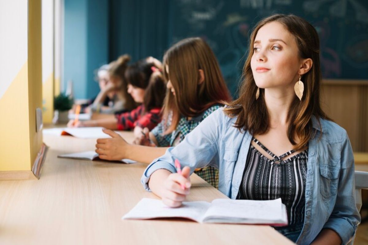 Uma aluna na sala de aula olha para fora pela janela. Ela está escrevendo no caderno. Ao fundo é possível ver outros colegas de classe 