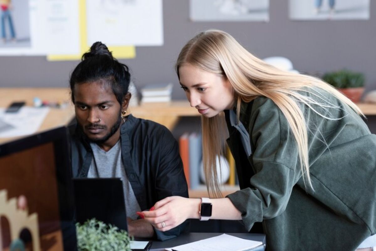 Dois alunos olham atentamente para o notebook. Um aluno está sentado, e sua amiga olha por cima dos seus ombros e ponta para a tela vendo se tem redação na prova da ETEC