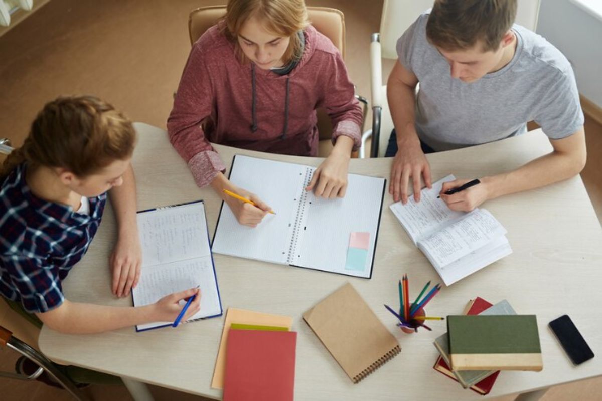 Três alunos estudando [vista de cima]. Eles usam cadernos e escrevem neles como teste para a redação