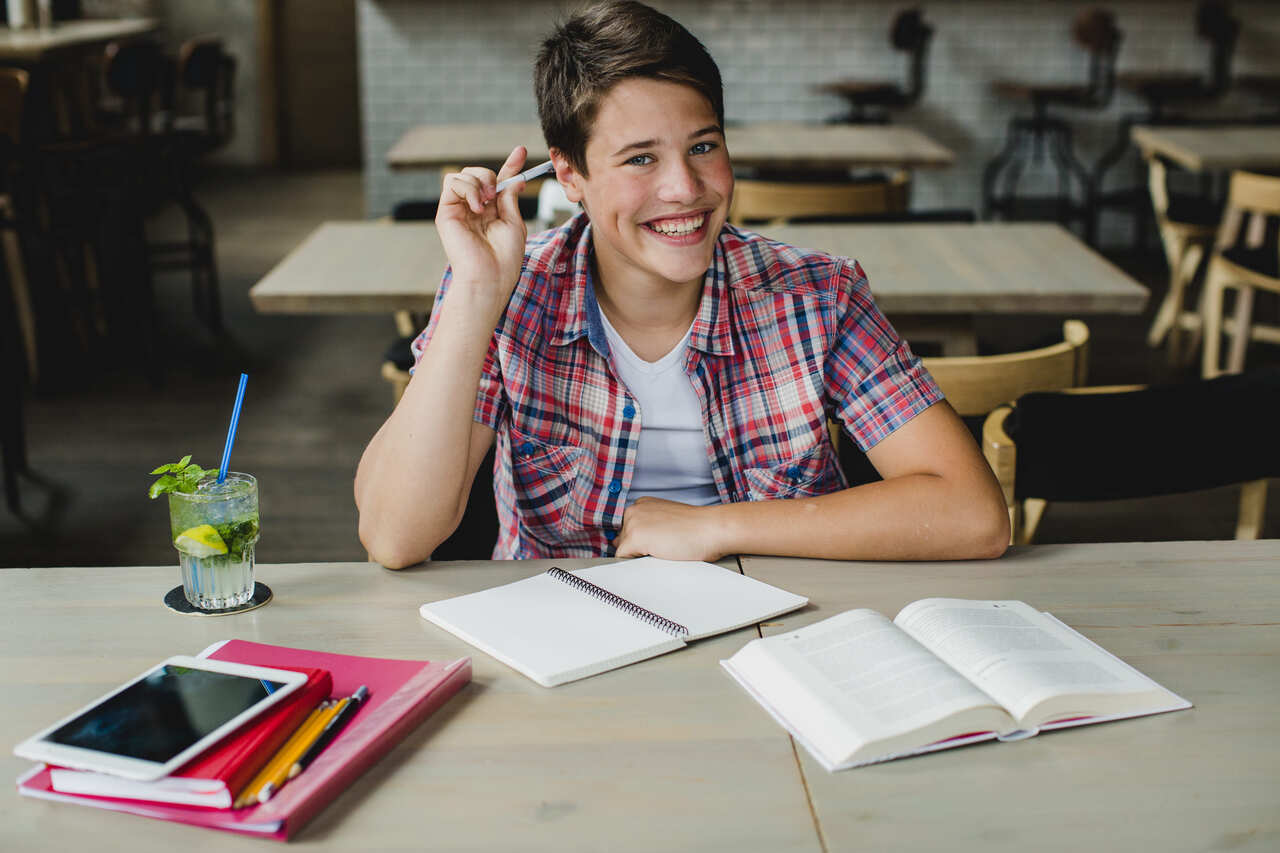 Jovem posa sorridente sentado em mesa diante materiais de estudo 