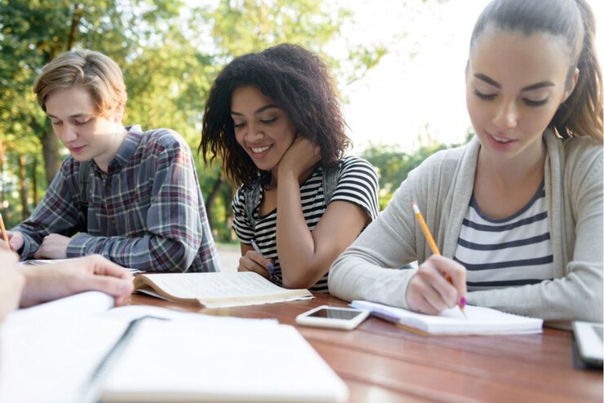 Três alunos em uma mesa. Uma estudante escreve em seu caderno, a outra lê um livro e seu amigo fala o que estudar para o Vestibulinho ETEC 2025