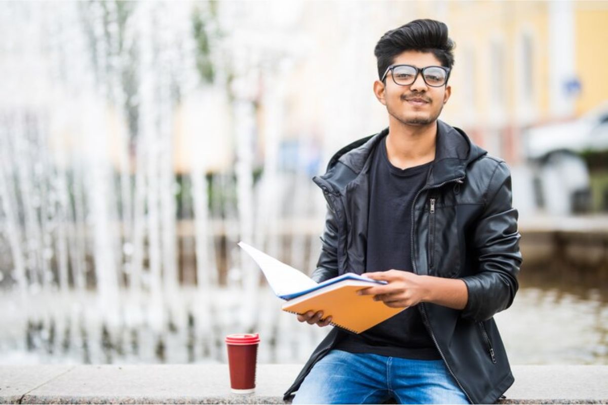 Um estudante sentado em frente a uma fonte de água na praça. Ele está segurando um caderno aberto