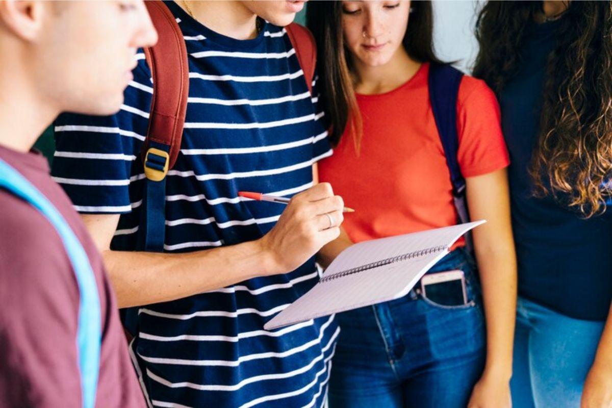 Três alunos em pé na sala de aula. Um (no meio) segura um caderno e outros dois (ao lado) olham