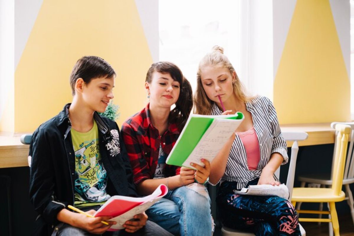 Estudantes em uma sala de aula. Três alunos leem uma apostila. Eles querem saber o que estudar para o Vestibulinho ETEC 2025